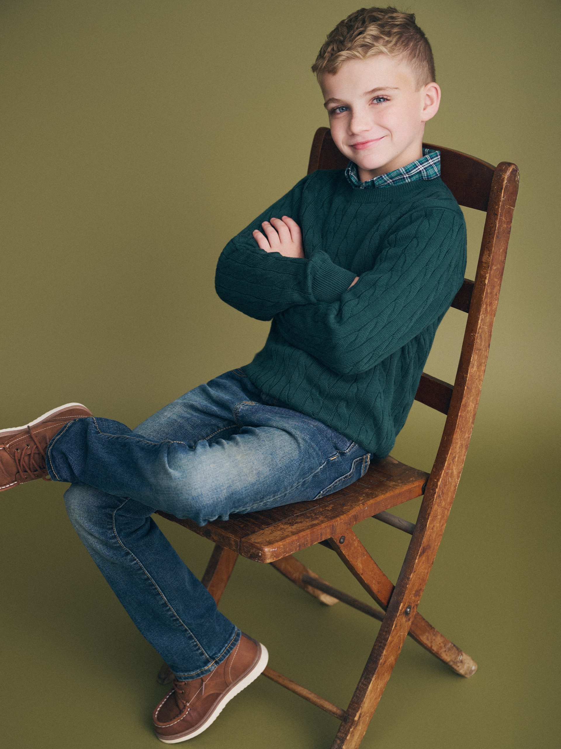Declan_11 Child in a green sweater and jeans sitting sideways on a wooden chair with arms crossed.